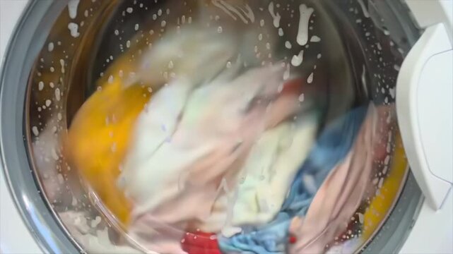 Front view through washing machine glass door as drum spins rapidly, creating blurred circular motion of colorful fabrics, highlighting household routine, cleaning process and dynamic rotation pattern