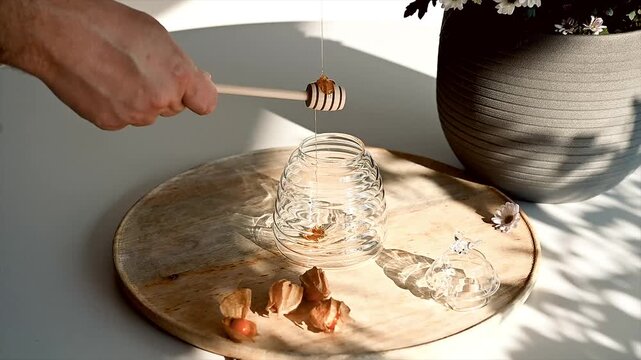 A hand holds a wooden honey dipper above an empty glass beehive jar on a round wooden board with physalis and a glass bee lid in bright daylight.