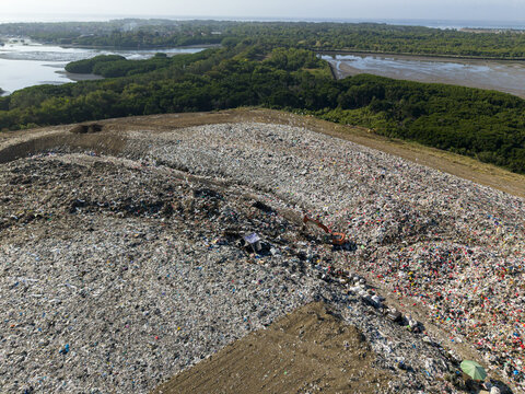 Aerial view of TPA Suwung landfill with piles of waste, heavy machinery operating on site, and surrounding mangrove forests in Kecamatan Denpasar Selatan, Bali, Indonesia.