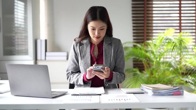 Professional business woman working at her office desk using a calculator to check finances and holding her smartphone while reviewing charts and documents on her desk