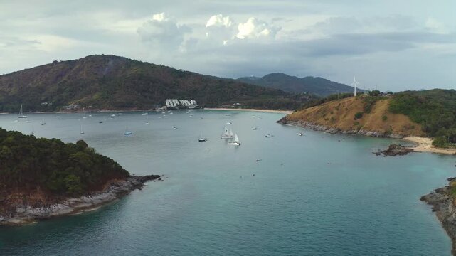 Panoramic aerial view capturing Nai Harn Beach in Phuket, Thailand. Numerous sailboats anchored in sea bay. People swimming and enjoying the tropical ocean shore. Zoom out drone flight landscape