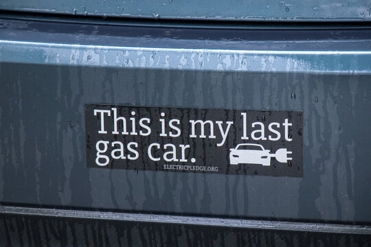A black This is my last gas car bumper sticker with an electric car icon and electricpledge.org affixed to a rain-speckled teal vehicle promoting EV adoption