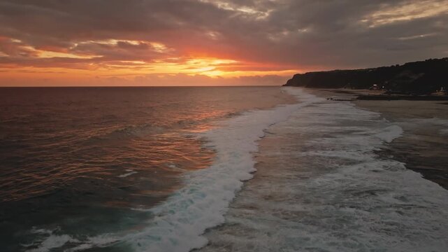 Melasti Beach during a warm sunset, foamy ocean waves breaking gently along the dark sandy shore and reflecting the orange sky, creating a serene tropical coastal landscape. Aerial view drone flight