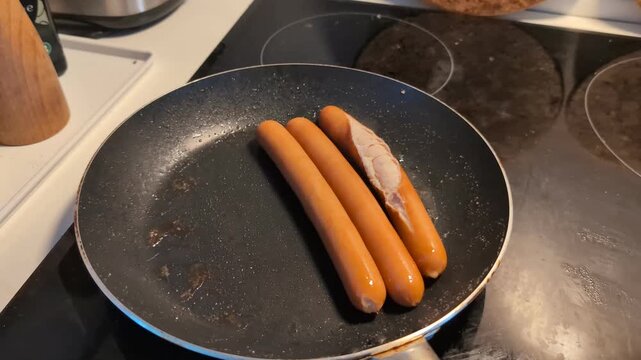 Frying three sausages in a hot non stick pan on an induction stove while one of them bursts open from the heat, revealing the meat inside during the cooking process