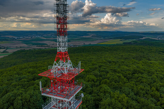 Aerial view of a red and white telecommunications tower rising above a dense forest with rolling hills in the background under a dramatic sky in Strazovice, South Moravian Region, Czechia.