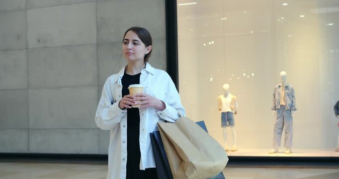 Young white woman holding takeaway coffee. Pauses outside fashion storefront, glancing at mannequins, carrying large paper bags, soft mall lighting and modern concrete wall create calm retail