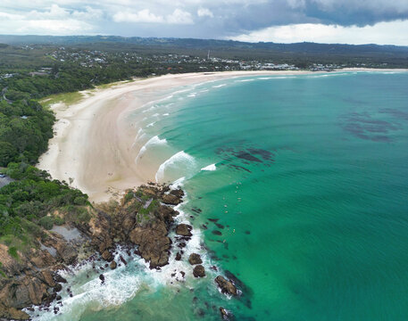 Aerial view of the coastline at Byron Bay with turquoise waves breaking on the sandy beach, rocky outcrops, and lush greenery in Byron Bay, New South Wales, Australia.