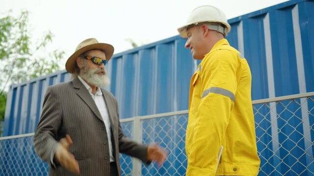 Senior caucasian business manager shaking hands with a logistics engineer in a yellow safety jacket and hard hat. Successful partnership deal at an industrial shipping container yard terminal outdoors