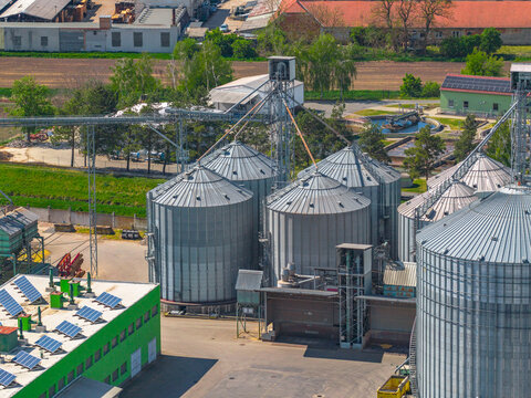 Aerial view of a grain storage facility featuring large metal silos, a green building with rooftop solar panels, and industrial equipment in Slavkov u Brna, South Moravian Region, Czechia.