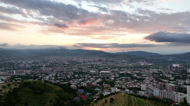 A high-angle aerial panoramic view overlooking the city of Tbilisi, Georgia, during a beautiful sunset. The expansive cityscape is nestled between rolling green hills in the foreground