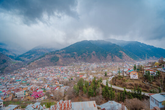 Aerial view of Bhaderwah town nestled in the mountains with colorful rooftops, winding roads, and dense forest slopes under a cloudy sky in Jammu, Jammu, India.