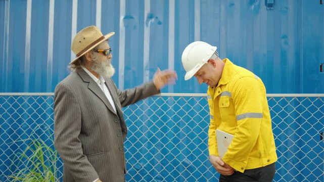 Senior businessman and young construction engineer shaking hands, talking and smiling in front of blue shipping container. Successful partnership, agreement, teamwork, and project management concept.