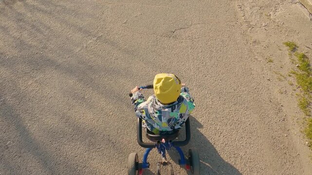 Overhead view of child on tricycle riding on paved path. Bright yellow hat colorful jacket. Warm sunlight casts long shadows. Casual outdoor activity