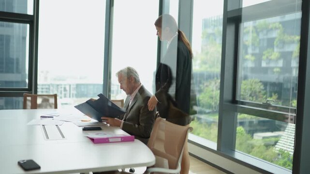 Female employee delivering business document to senior executive during corporate meeting before leaving office while experienced manager continues discussion with colleague in modern workplace