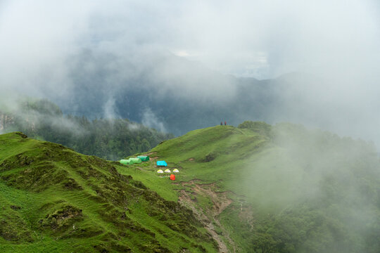 Aerial view of Ali Bugyal featuring colorful camping tents nestled on lush green mountain meadows surrounded by misty ridges in Chamoli, Uttarakhand, India.