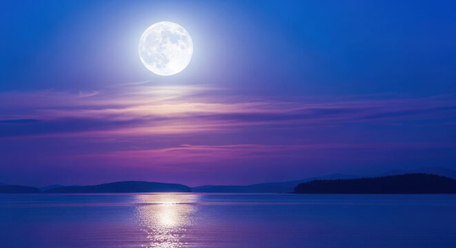 Large full moon shining brightly over calm water surface during quiet blue evening with soft clouds and reflection glimmering across peaceful sea.