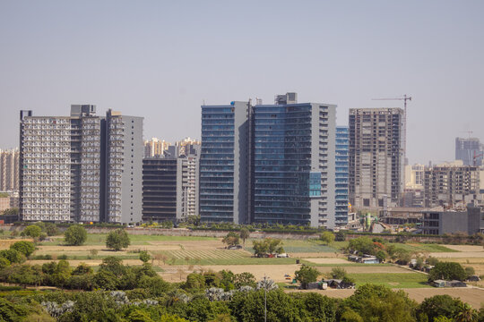 Aerial view of modern high-rise apartment buildings and construction sites rising above green agricultural fields and trees in the urban landscape of Noida, Uttar Pradesh, India.