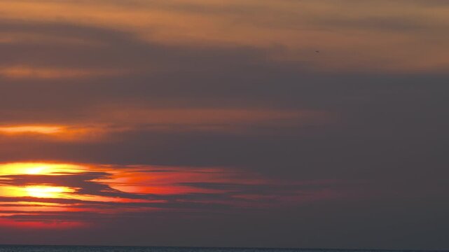 Distant airplane flying through dark clouds during dramatic red sunset over calm ocean, wide shot