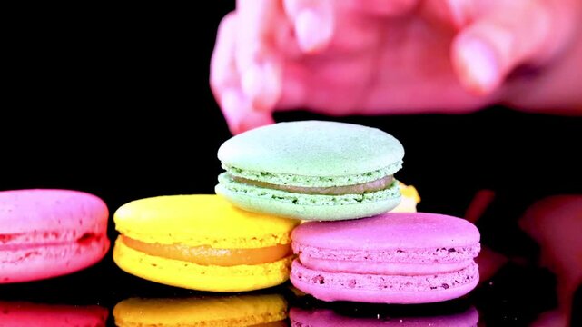 Close up shot of a hand stacking colorful French macarons including pink yellow and green flavors against a solid black background