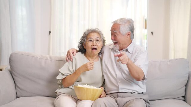 Happy Asian family senior couple relaxing on sofa watching television together at home, elderly husband and wife enjoy popcorn and leisure time, retirement bonding lifestyle and aging gracefully