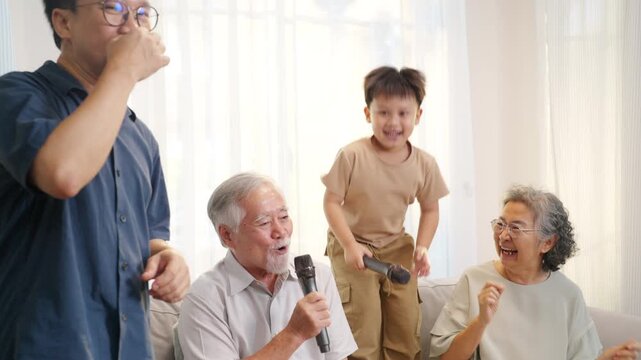 Happy multi-generation Asian family singing karaoke together at home. Grandparents, parent and young child enjoy music activity in living room, family bonding, retirement lifestyle joyful leisure time