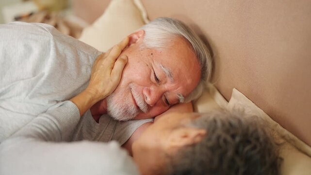 Asian senior couple sharing affectionate moment in bed at home. Elderly husband gently touching wife face during wake up in morning, loving relationship and retirement lifestyle with aging gracefully