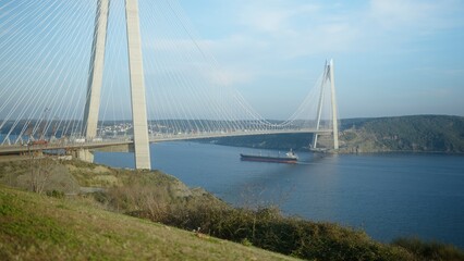 Obraz premium Panoramic view of a large cable-stayed bridge over a blue river with a cargo ship passing beneath on a clear day