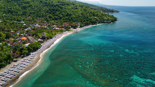 Aerial view of Bintang Beach Villa with traditional outrigger boats on the dark sand beach, turquoise coral reefs, and lush green hills in Kecamatan Abang, Bali, Indonesia.