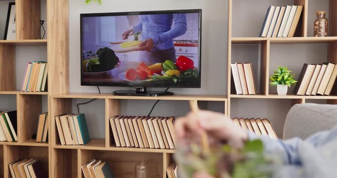 TV cooking tutorial vegetables man salad watching. The screen in a living room shows hands slicing fresh produce as a viewer is eating a bowl of greens. Healthy learning for home kitchens.