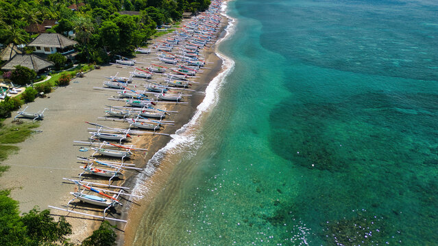 Aerial view of Bintang Beach Villa and traditional jukung boats lined up on the dark sand beach along the turquoise waters of the Bali Sea in Kecamatan Abang, Bali, Indonesia.
