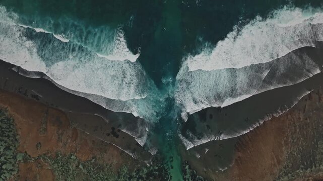 Drone flight over white ocean waves breaking on the rugged coastline of Melasti Beach in Bali, Indonesia, with a vibrant blue channel leading inland. Aerial top down view