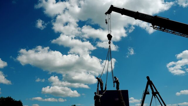 Silhouettes of riggers attaching lifting slings to heavy industrial cargo before crane hoisting. Construction, heavy lifting, engineering and industrial installation.