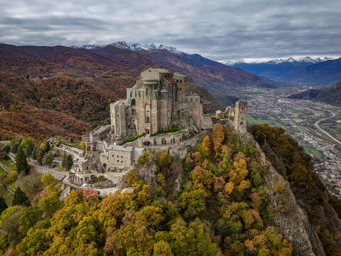Aerial view of Sacra di San Michele perched on a rocky peak surrounded by autumn forests with vibrant foliage and distant snow-capped mountains, Sant'Ambrogio di Torino, Piemonte, Italy.
