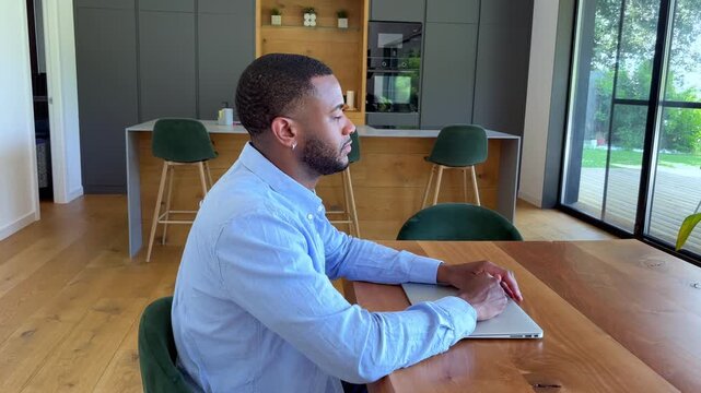 A young Black man sits thoughtfully at a stylish wooden table in a modern kitchen, with a closed laptop in front of him. He gazes off-screen, conveying patience or contemplation in a contemporary home