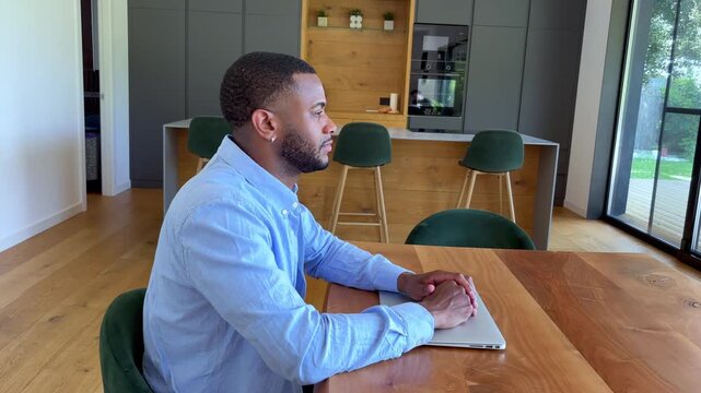 A focused young man sits at a modern dining table with a closed laptop, deep in thought or patiently waiting. His expression suggests anticipation or contemplation in a contemporary home setting.