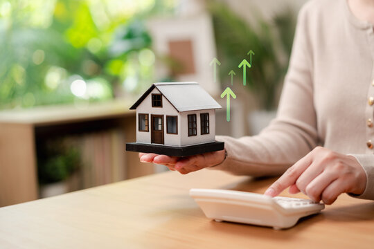 Rising property value and mortgage interest rate concept. Woman calculating home loan cost with miniature house and up arrows.