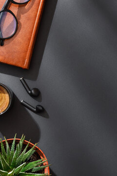 Leather notebook with wireless earbuds, glasses, black coffee espresso cup and succulent plant on black background in dramatic soft lighting. Modern dark workspace. Flat lay with copy space.
