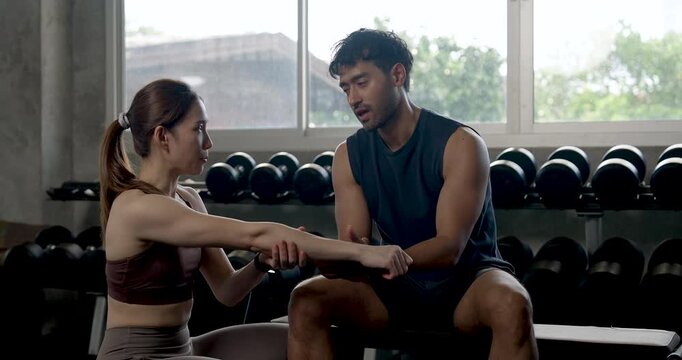 Young Asian couple practices partner training in modern gym, woman kneeling by seated man to guide wrist grip and dumbbell form, building support, motivation, wellness, healthy workout teamwork