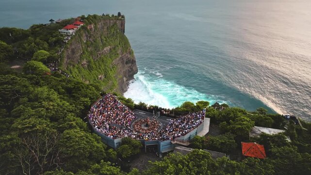 Crowds of tourists are watching a traditional Balinese Kecak fire dance in an open-air amphitheater at Uluwatu Temple, perched on a cliff edge overlooking the Indian Ocean. Aerial view drone shot
