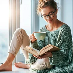 Woman reads a book and drinks coffee while sitting by a window on a sunny afternoon © alphaspirit