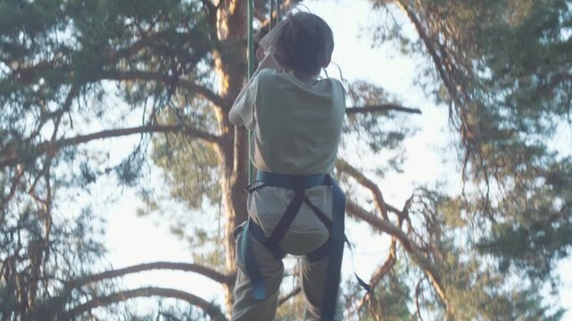 A happy boy wearing a safety harness rides a fast zipline down a cable in an adventure rope park. The child smiles joyfully and flies above the ground against the background of tall pine trees
