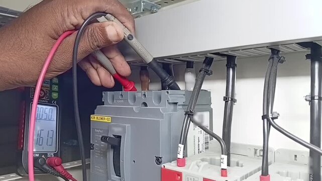 Electrician using digital multimeter probes to check voltage between L1 and L3 phases on industrial MCCB breaker inside electrical panel
