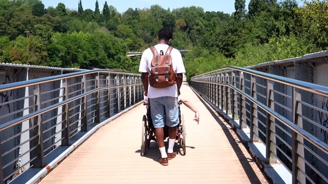 Rear view of a young caregiver assisting a disabled person in a wheelchair, crossing a pedestrian bridge surrounded by lush green trees on a sunny day and promoting inclusion and accessibility