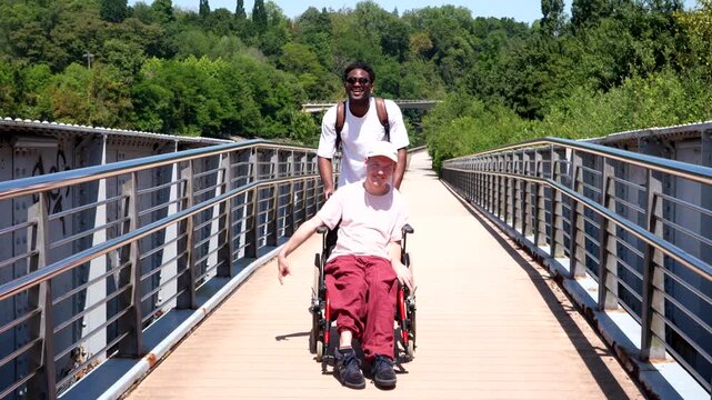 Happy african american man pushing his caucasian friend in a wheelchair along a sunny pedestrian bridge, demonstrating concepts of friendship, support, inclusion, and enjoying outdoor life