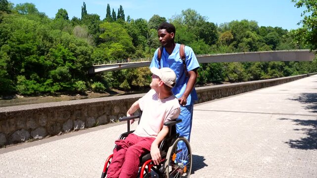 Young black male caregiver pushing a disabled man in a wheelchair along a beautiful riverwalk in a city park, providing compassionate assistance and support on a sunny day outdoors