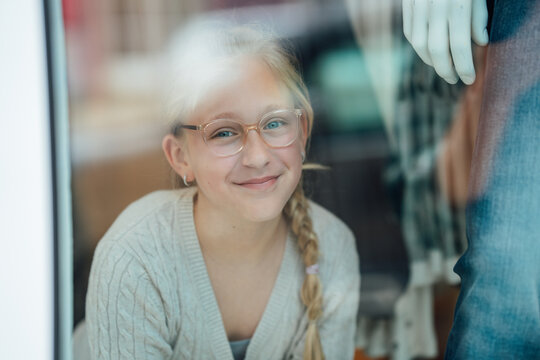 Smiling child with glasses standing behind a fashion store window looking outside at the street enjoying an urban shopping experience.