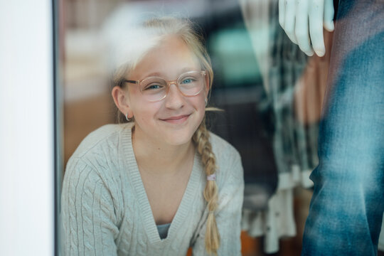 Happy child wearing glasses looking through the fashion store window with curiosity while watching the busy street and city life outside.