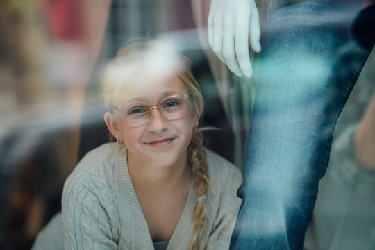 Happy young child in glasses looks through a large fashion store window toward the street capturing a joyful urban lifestyle moment.