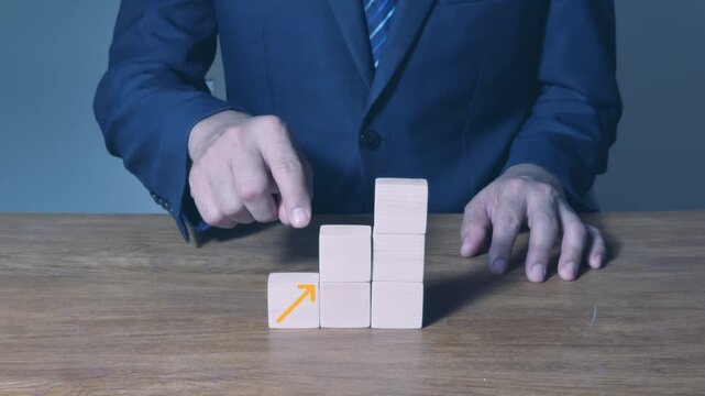 Close-up of a businessman touching a wooden block with a target and arrow icon, completing a step chart with rising arrow graphics for goal achievement, growth, and success concept.