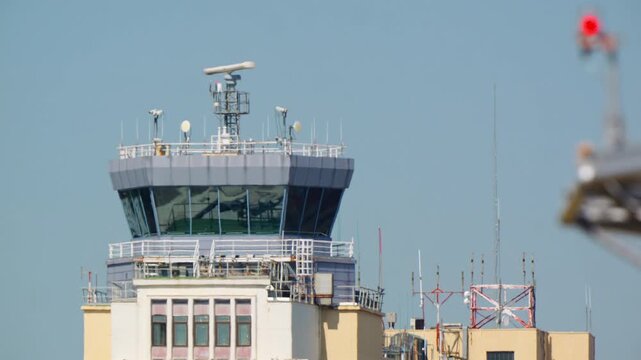 Airport control tower with rotating radar dish antenna on top, surrounded by various communication antennas and transmission towers, clear blue sky background with distant runway lights visible
